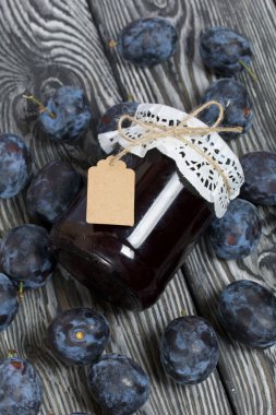 Bir kavanoz erik reçeli. Üzerinde bir yazıt etiketi var. Erikler yakınlarda dağılmış. Çam ağaçlarında.