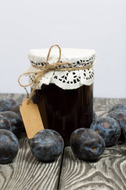 Bir kavanoz erik reçeli. Üzerinde bir yazıt etiketi var. Erikler yakınlarda dağılmış. Çam ağaçlarında.