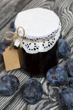 Bir kavanoz erik reçeli. Üzerinde bir yazıt etiketi var. Erikler yakınlarda dağılmış. Çam ağaçlarında.