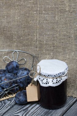 Bir kavanoz erik reçeli. Üzerinde bir yazıt etiketi var. Sepetin içindeki eriklerin yanında. Çam ağaçlarında.