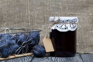 Bir kavanoz erik reçeli. Üzerinde bir yazıt etiketi var. Sepetin içindeki eriklerin yanında. Çam ağaçlarında.