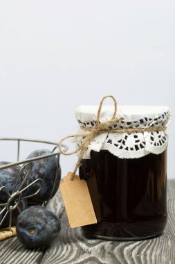 Bir kavanoz erik reçeli. Üzerinde bir yazıt etiketi var. Sepetin içindeki eriklerin yanında. Çam ağaçlarında.