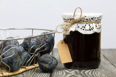 Bir kavanoz erik reçeli. Üzerinde bir yazıt etiketi var. Sepetin içindeki eriklerin yanında. Çam ağaçlarında.