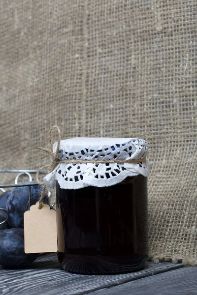 Bir kavanoz erik reçeli. Üzerinde bir yazıt etiketi var. Sepetin içindeki eriklerin yanında. Çam ağaçlarında.