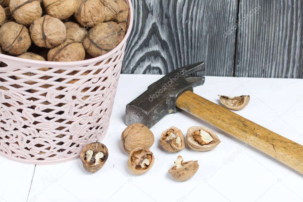 A basket with walnuts. Nearby is a hammer and chopped nuts. On a white surface.