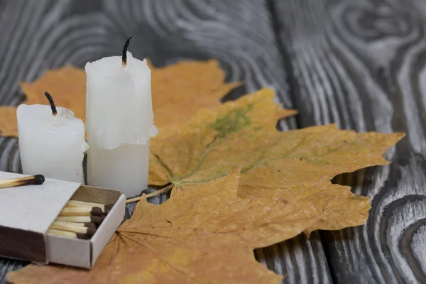 Two candlesticks on brushed pine boards. Nearby are boxes of matches and dried maple leaves.