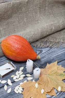 Portakal kabağı, tohumlar ve mum koçanları. Yakınlarda kibrit kutuları ve kurumuş akçaağaç yaprakları var. Çam ağacından yapılmış tahtalarda siyah beyaz boyanmış..