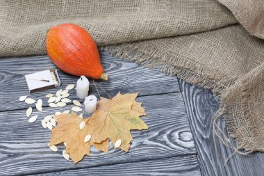Portakal kabağı, tohumlar ve mum koçanları. Yakınlarda kibrit kutuları ve kurumuş akçaağaç yaprakları var. Çam ağacından yapılmış tahtalarda siyah beyaz boyanmış..