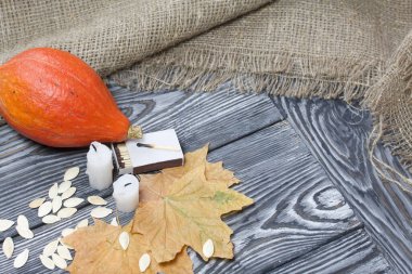 Portakal kabağı, tohumlar ve mum koçanları. Yakınlarda kibrit kutuları ve kurumuş akçaağaç yaprakları var. Çam ağacından yapılmış tahtalarda siyah beyaz boyanmış..