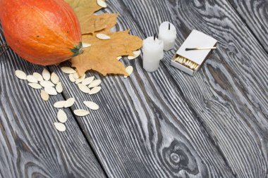 Portakal kabağı, tohumlar ve mum koçanları. Yakınlarda kibrit kutuları ve kurumuş akçaağaç yaprakları var. Çam ağacından yapılmış tahtalarda siyah beyaz boyanmış..