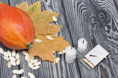 Portakal kabağı, tohumlar ve mum koçanları. Yakınlarda kibrit kutuları ve kurumuş akçaağaç yaprakları var. Çam ağacından yapılmış tahtalarda siyah beyaz boyanmış..