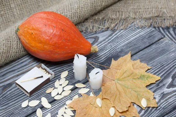 Portakal kabağı, tohumlar ve mum koçanları. Yakınlarda kibrit kutuları ve kurumuş akçaağaç yaprakları var. Çam ağacından yapılmış tahtalarda siyah beyaz boyanmış..