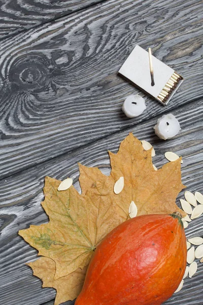 Portakal kabağı, tohumlar ve mum koçanları. Yakınlarda kibrit kutuları ve kurumuş akçaağaç yaprakları var. Çam ağacından yapılmış tahtalarda siyah beyaz boyanmış..