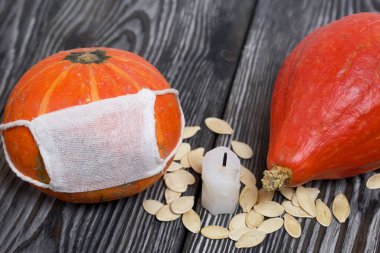 Tıbbi maskeli turuncu balkabağı. Yakınlarda bir balkabağı daha var, bir mum var ve kabak tohumları etrafa saçılmış. Siyah beyaz renklerle boyanmış çam ağaçlarının arka planında.