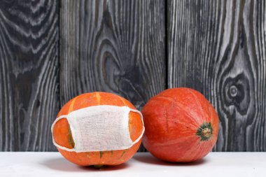 Tıbbi maskeli turuncu balkabağı. Yakınlarda maskesiz bir balkabağı var. Çam ağaçlarının arka planında, siyah beyaz boyanmış..