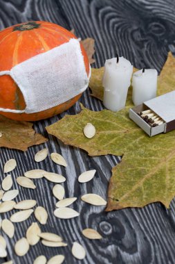 Tıbbi maskeli turuncu balkabağı. Yakınlarda mumlar, kabak tohumları ve kurumuş akçaağaç yaprakları var. Siyah beyaz renklerle boyanmış çam ağaçlarının arka planında.