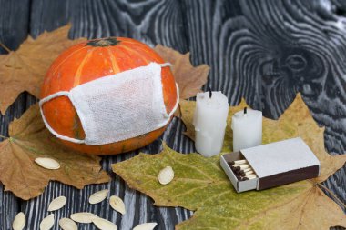 Tıbbi maskeli turuncu balkabağı. Yakınlarda mumlar, kabak tohumları ve kurumuş akçaağaç yaprakları var. Siyah beyaz renklerle boyanmış çam ağaçlarının arka planında.