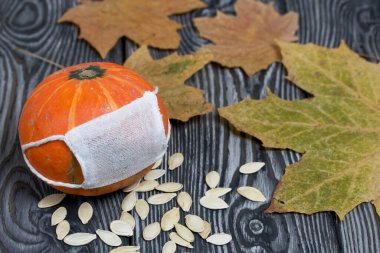 Tıbbi maskeli turuncu balkabağı. Balkabağı tohumu ve kurumuş akçaağaç yaprakları etrafa saçılmış. Siyah beyaz renklerle boyanmış çam ağaçlarının arka planında.