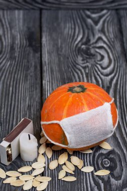 Tıbbi maskeli turuncu balkabağı. Yakınlarda bir mum var ve balkabağı tohumları etrafa saçılmış. Siyah beyaz renklerle boyanmış çam ağaçlarının arka planında.