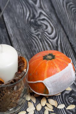 Tıbbi maskeli turuncu balkabağı. Yakınlarda bir mum var ve balkabağı tohumları etrafa saçılmış. Siyah beyaz renklerle boyanmış çam ağaçlarının arka planında.