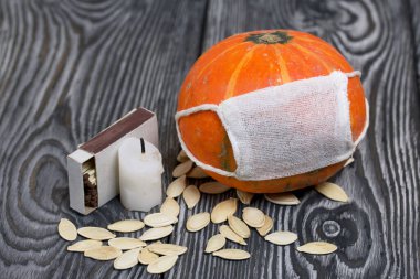 Tıbbi maskeli turuncu balkabağı. Yakınlarda bir mum var ve balkabağı tohumları etrafa saçılmış. Siyah beyaz renklerle boyanmış çam ağaçlarının arka planında.