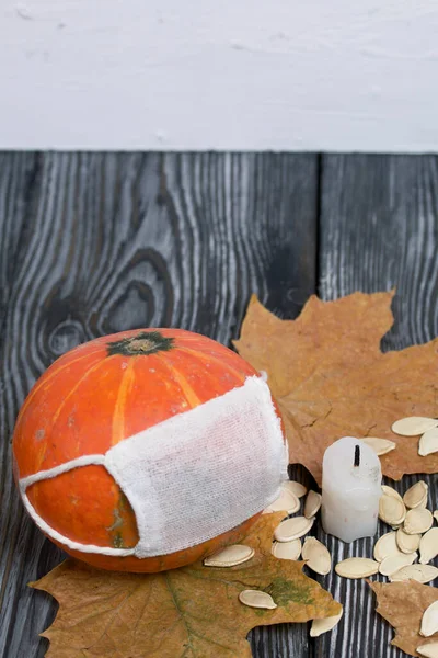 Tıbbi maskeli turuncu balkabağı. Yakınlarda bir mum, kabak tohumları ve kurumuş akçaağaç yaprakları var. Siyah beyaz renklerle boyanmış çam ağaçlarının arka planında.