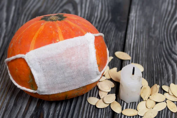 Tıbbi maskeli turuncu balkabağı. Yakınlarda bir mum var ve balkabağı tohumları etrafa saçılmış. Siyah beyaz renklerle boyanmış çam ağaçlarının arka planında.