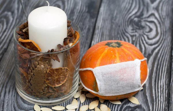 Tıbbi maskeli turuncu balkabağı. Yakınlarda bir mum var ve balkabağı tohumları etrafa saçılmış. Siyah beyaz renklerle boyanmış çam ağaçlarının arka planında.