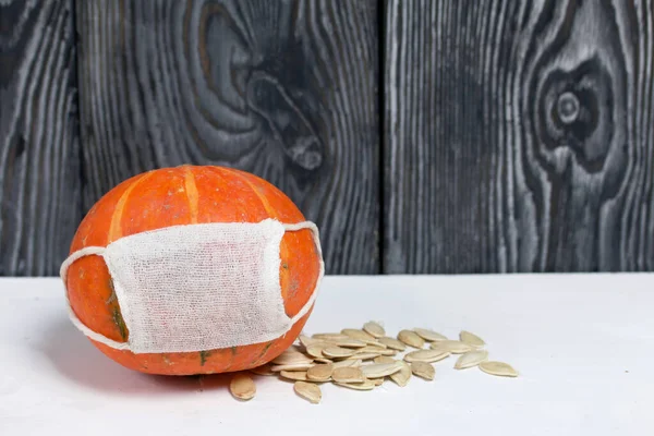 Orange pumpkin with a medical mask. Pumpkin seeds are scattered nearby. Against the background of brushed pine boards painted in black and white.