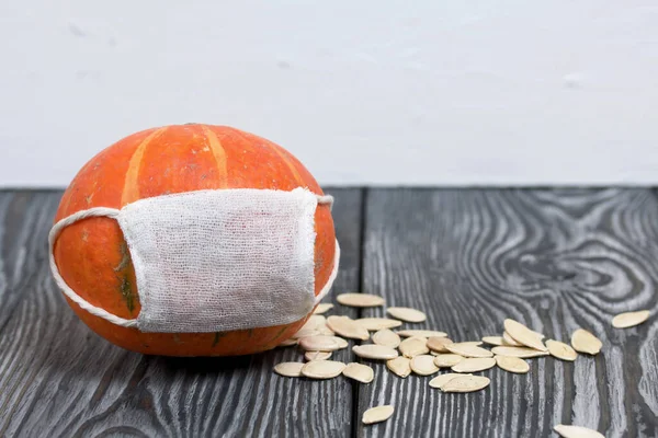 Tıbbi maskeli turuncu balkabağı. Balkabağı tohumları yakınlarda serpilmiş. Siyah beyaz renklerle boyanmış çam ağaçlarının arka planında.