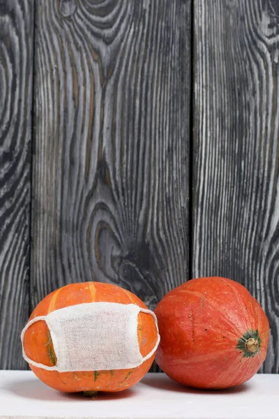Tıbbi maskeli turuncu balkabağı. Yakınlarda maskesiz bir balkabağı var. Çam ağaçlarının arka planında, siyah beyaz boyanmış..
