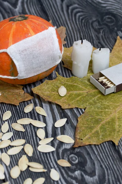 Tıbbi maskeli turuncu balkabağı. Yakınlarda mumlar, kabak tohumları ve kurumuş akçaağaç yaprakları var. Siyah beyaz renklerle boyanmış çam ağaçlarının arka planında.
