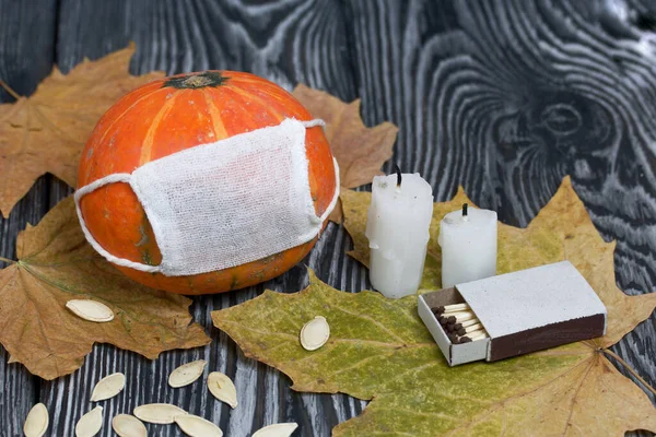 Tıbbi maskeli turuncu balkabağı. Yakınlarda mumlar, kabak tohumları ve kurumuş akçaağaç yaprakları var. Siyah beyaz renklerle boyanmış çam ağaçlarının arka planında.