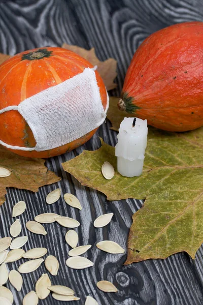 Tıbbi maskeli turuncu balkabağı. Yakınlarda bir balkabağı ve bir mum daha var. Balkabağı tohumları ve kurumuş akçaağaç yaprakları dağılmış. Siyah beyaz renklerle boyanmış çam ağaçlarının arka planında.