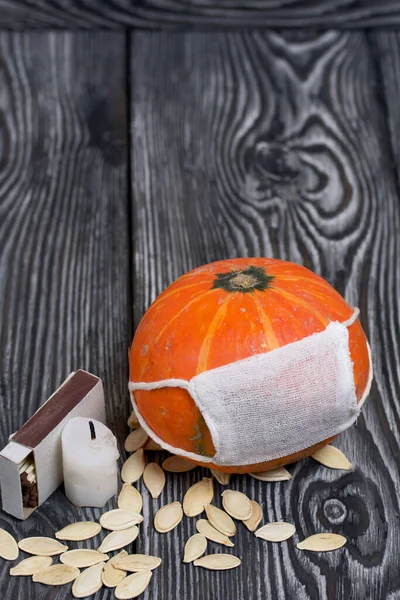 Tıbbi maskeli turuncu balkabağı. Yakınlarda bir mum var ve balkabağı tohumları etrafa saçılmış. Siyah beyaz renklerle boyanmış çam ağaçlarının arka planında.