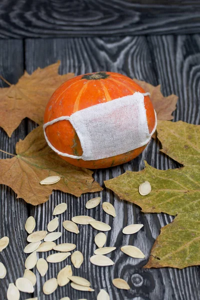 Orange pumpkin with a medical mask. Pumpkin seeds and dried maple leaves are scattered nearby. Against the background of brushed pine boards painted in black and white.