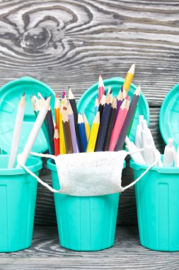 Renkli kalemler, tükenmez kalemler. Tıbbi maske takıyorlar. Çöp tenekesi şeklinde bir kalem kutusu. Çam ağacından yapılmış tahtalarda siyah beyaz boyanmış..