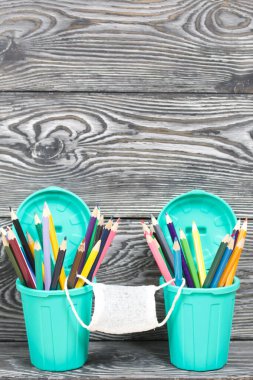 Kalem tutacaklarında renkli kalemler. Tıbbi maske takıyorlar. Çöp tenekesi şeklinde bir kalem kutusu. Çam ağacından yapılmış tahtalarda siyah beyaz boyanmış..