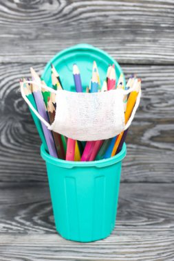 Kalem tutacağındaki renkli kalemler. Tıbbi maske takıyorlar. Çöp tenekesi şeklinde bir kalem kutusu. Çam ağacından yapılmış tahtalarda siyah beyaz boyanmış..