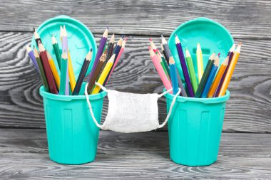 Kalem tutacaklarında renkli kalemler. Tıbbi maske takıyorlar. Çöp tenekesi şeklinde bir kalem kutusu. Çam ağacından yapılmış tahtalarda siyah beyaz boyanmış..