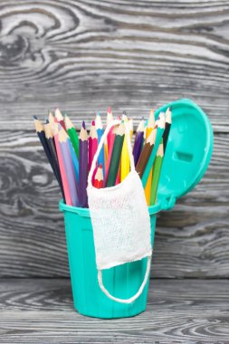 Kalem tutacağındaki renkli kalemler. Tıbbi maske takıyorlar. Çöp tenekesi şeklinde bir kalem kutusu. Çam ağacından yapılmış tahtalarda siyah beyaz boyanmış..