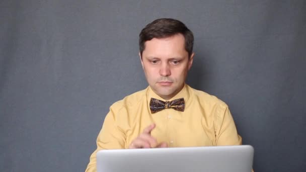 Мужчина в жёлтой рубашке и галстуке-бабочке сидит за ноутбуком. Общается с собеседником по видеосвязи и надевает медицинскую маску. Снимок сделан на сером фоне. — стоковое видео