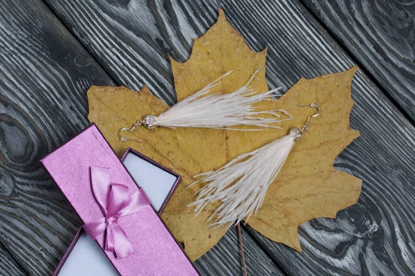 Ostrich feather brush earrings and a case for them. Nearby are dried maple leaves. They lie on pine boards painted in black and white. Autumn gifts for loved ones.