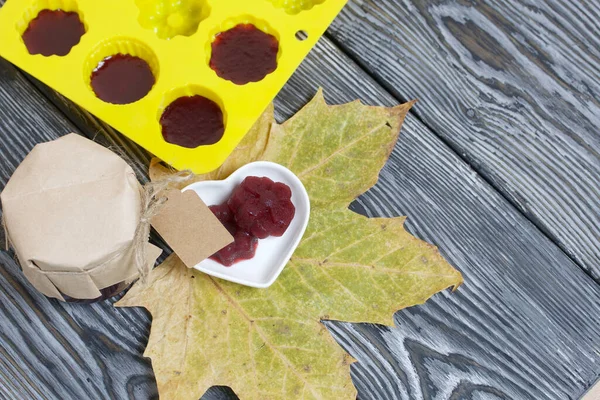 A jar of cherry jam with a label for an inscription and a saucer of cherry marmalade. A silicone mold with marmalade is visible. Dried maple leaves all around. On pine planks painted black and white.