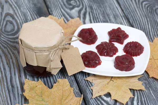 A jar of cherry jam with a label for an inscription and a saucer of cherry marmalade. Dried maple leaves all around. On pine planks painted black and white.