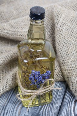 Bir şişe lavanta tentürü. Şişeye bir demet lavanta bağlanmış. Yakınlarda kalın keten kumaş var. Siyah-beyaz boyanmış çam ağaçlarında..