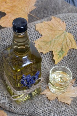 Bir şişe lavanta tentürü. Şişeye bir demet lavanta bağlanmış. Yakınlarda bir bardak içki ve kurumuş akçaağaç yaprakları var. Kaba ketenler üzerinde. Yakın çekim..