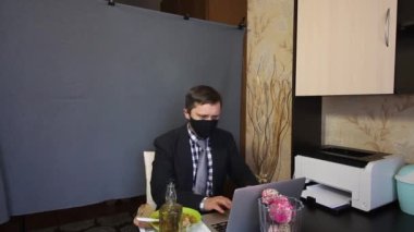 Video görüşmesi sırasında yemek yemek. Takım elbiseli ve yüzünde koruyucu maske olan bir adam dizüstü bilgisayarının başına oturup video iletişimi kuruyor. Onun yanındaki tepside yiyecek ve alkol var. Gri bir c