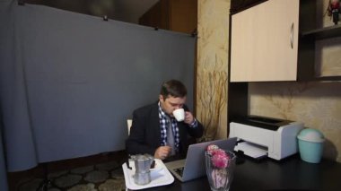 Video görüşmesi sırasında yemek yemek. Takım elbiseli bir adam video iletişimiyle konuşuyor. İletişim kuruyor, kahve içiyor. Tepsinin yanında bir kahve makinesi ve bir kek var..