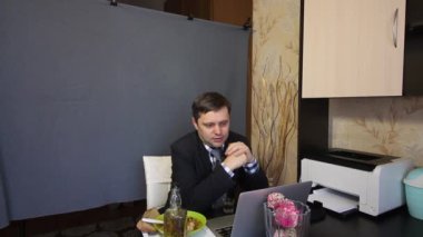Video görüşmesi sırasında yemek yemek. Takım elbiseli bir adam video iletişimiyle konuşuyor. Bardağa alkol ve içki doldurur. Onun yanındaki tepside yemek var..
